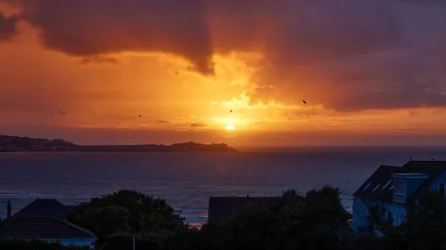 St Ives Sunset This landscape photograph captures a vivid sunset over the sea at St Ives, located in Cornwall, England, United Kingdom. The image was taken in the evening during the summer season, as evidenced by the low sun descending towards the horizon and the warm light illuminating the sky and clouds. The foreground features residential buildings and trees, while the coastline of St Ives is visible across the water. The dramatic colors and rays in the sky showcase the typical beauty of a Cornwall summer sunset over the sea, with a few birds silhouetted against the sky, enhancing the tranquil scene.